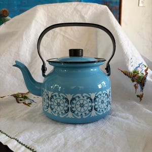 May include: A light blue enamel teapot with a black handle and lid. The teapot features a white and blue decorative pattern around the body. The teapot is sitting on a white cloth.