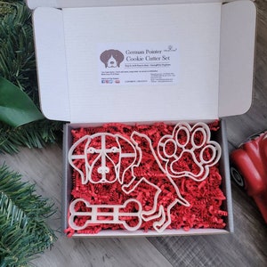 May include: A white cardboard box containing a German Pointer cookie cutter set. The set includes cutters shaped like a dog, a paw print, and a bone. The box is filled with red paper shreds. The box has text that reads "German Pointer Cookie Cutter Set".