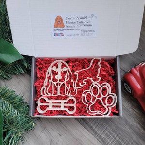 May include: A set of four white plastic cookie cutters in a red and white box. The cutters are shaped like a cocker spaniel dog, a paw print, a bone, and a side view of a cocker spaniel. The box is filled with red shredded paper. The text "Cocker Spaniel Cookie Cutter Set" is printed on the box.