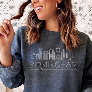 以下が含まれることがあります： 白いアウトラインのバーミンガムの街並みと、その下に「BIRMINGHAM」という文字がプリントされた濃い青色のスウェットシャツです。