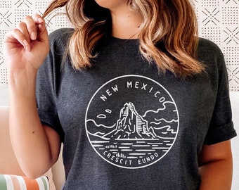 May include: A dark gray t-shirt with a white graphic design of a mountain range and the text "New Mexico Crescit Eundo".