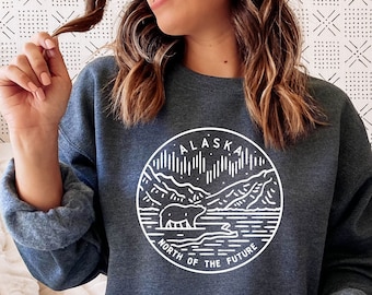 Könnte beinhalten: Ein dunkelblauer Pullover mit einem weißen Grafikdesign eines Bären und Bergen in einem Kreis. Der Text "Alaska North of the Future" befindet sich im Kreis.
