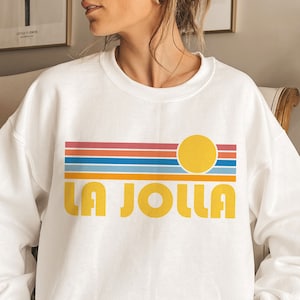 May include: White sweatshirt with a retro graphic design featuring a yellow sun, colorful stripes, and the text "LA JOLLA" in yellow.