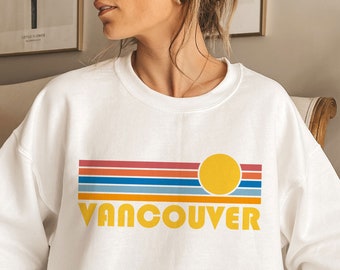 May include: White sweatshirt with a retro-style graphic design featuring a yellow sun, colorful stripes, and the word "VANCOUVER" in yellow letters.