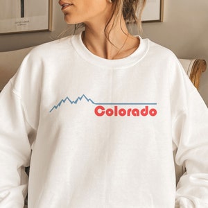 May include: White sweatshirt with a graphic of blue mountain peaks and the word "Colorado" in red. The sweatshirt has a crew neck and long sleeves. The design is simple and clean, perfect for casual wear.