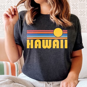 May include: A woman wearing a dark grey t-shirt with a retro sunset graphic and the word "HAWAII" in yellow letters. She is sitting on a couch with a striped pillow.