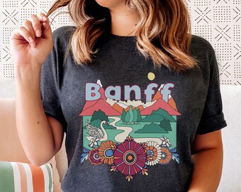Könnte beinhalten: Ein dunkelgraues T-Shirt mit einem Retro-Grafikdesign, das eine Berglandschaft, einen Fluss, einen Schwan und Blumen zeigt. Der Text "Banff" ist in Weiß auf das Shirt gedruckt.