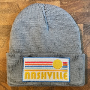 May include: A light gray beanie with a rectangular patch featuring a sun and horizontal stripes in red, orange, yellow, and blue. The word "NASHVILLE" is embroidered in yellow. The beanie is made of a knitted material.