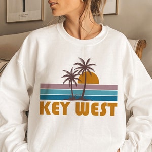 Puede incluir: Sudadera blanca con un gráfico de puesta de sol retro con palmeras y el texto "KEY WEST" en una fuente naranja en negrita.