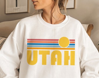 May include: White sweatshirt with a retro-style graphic design featuring a yellow sun and the word "UTAH" in yellow letters. The graphic is set against a background of colourful stripes.