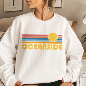 Könnte beinhalten: Weißer Pullover mit einem Retro-Sonnenuntergang-Motiv und dem Text "OCEANSIDE" in gelben Buchstaben.