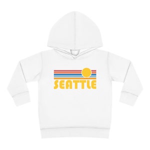 Pode incluir: Moletom branco com capuz para crian&ccedil;as pequenas com um gr&aacute;fico retr&ocirc; de um sol amarelo se pondo sobre um horizonte listrado azul, vermelho e branco. O texto "SEATTLE" &eacute; impresso em amarelo abaixo do gr&aacute;fico.