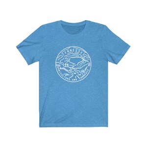 May include: A light blue t-shirt with a white graphic of the state seal of Tennessee. The seal features a mountain range, a river, and two figures standing on a hill. The text around the seal reads "Tennessee Agriculture and Commerce".