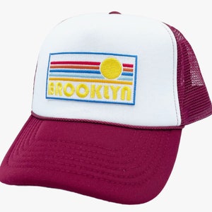 May include: A white and burgundy trucker hat with a retro-style embroidered patch that says "BROOKLYN" with a yellow sun and rainbow stripes.
