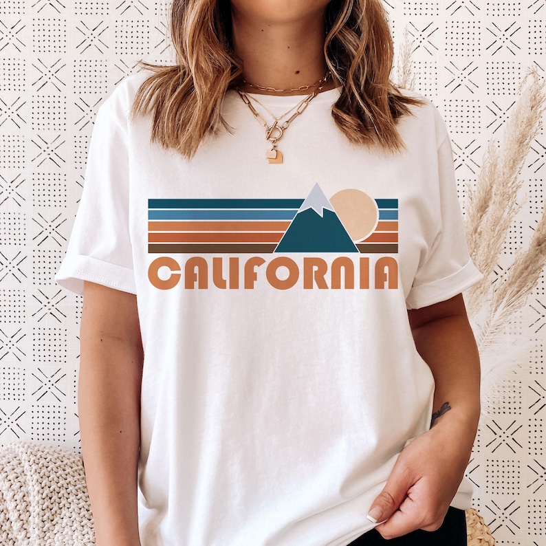 May include: White t-shirt with a retro graphic design featuring a mountain range, a sun, and the word "CALIFORNIA" in orange letters.