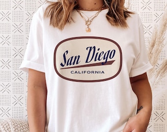 Könnte beinhalten: Weißes T-Shirt mit einem braunen ovalen Grafik, auf dem "San Diego, Kalifornien" in einer Vintage-Schriftart steht.