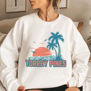 Puede incluir: Sudadera blanca con un gráfico de una puesta de sol, palmeras y olas del océano. El texto "Torrey Pines" está impreso debajo del gráfico.