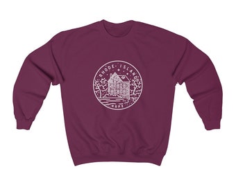 May include: A maroon sweatshirt featuring a white circular graphic with the words "RHODE ISLAND" and "HOPE" surrounding a detailed illustration of a house, trees, and water. The design is centered on the front of the crewneck.