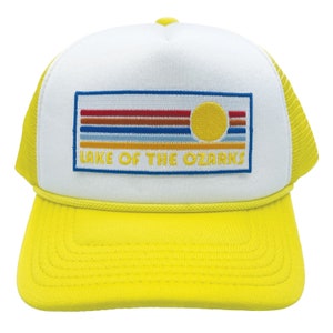 May include: A white and yellow trucker hat with a blue, red, and yellow embroidered patch that says "Lake of the Ozarks". The patch features a stylized sun and horizontal stripes.