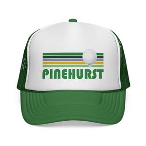 May include: Green and white trucker hat with a golf ball graphic and the word "PINEHURST" in green block letters. The hat features a green brim and mesh back, with a white front panel displaying the design.