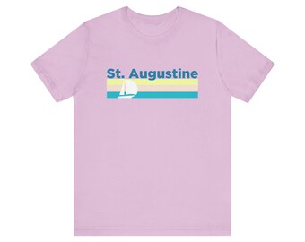 May include: A light purple t-shirt with a retro-style graphic design featuring the text "St. Augustine" and a sailboat silhouette against a yellow, blue, and white striped background.