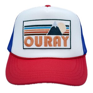 May include: A white and red trucker hat with a blue mesh back. The front of the hat features an embroidered patch with a mountain range, a sun, and the word "OURAY" in orange.