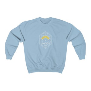 May include: Light blue sweatshirt with a white graphic of a mountain range with a sun rising behind it. The graphic is inside a hexagon with the text "ASPEN CO" below it.