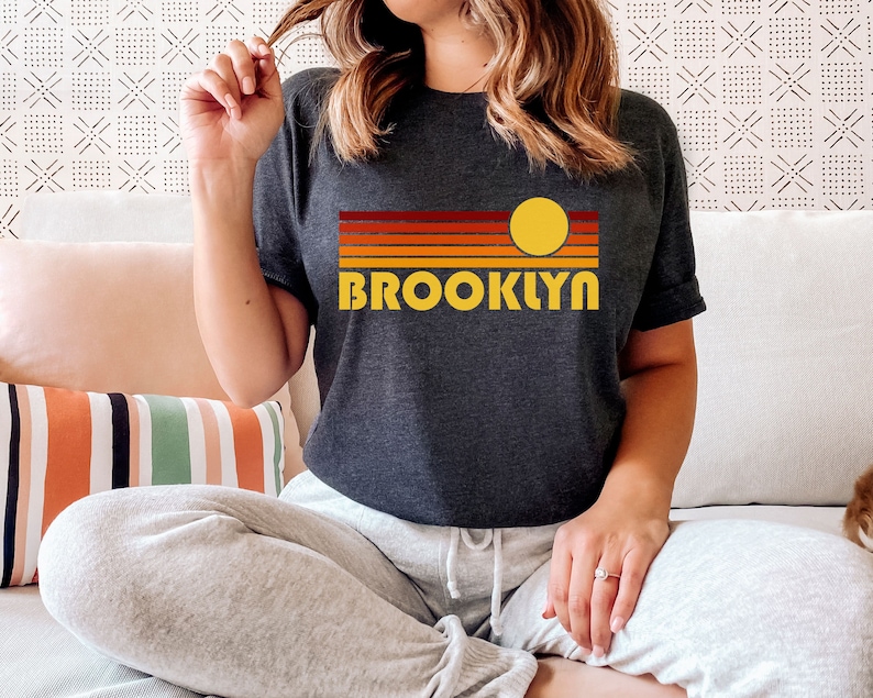 May include: A dark grey t-shirt with a retro sunset graphic and the word "BROOKLYN" in yellow lettering.