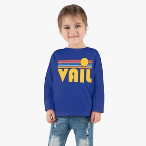 May include: A royal blue long-sleeved sweatshirt with a retro-style graphic design. The graphic features a yellow sun, blue, red, and orange stripes, and the word "VAIL" in yellow letters.