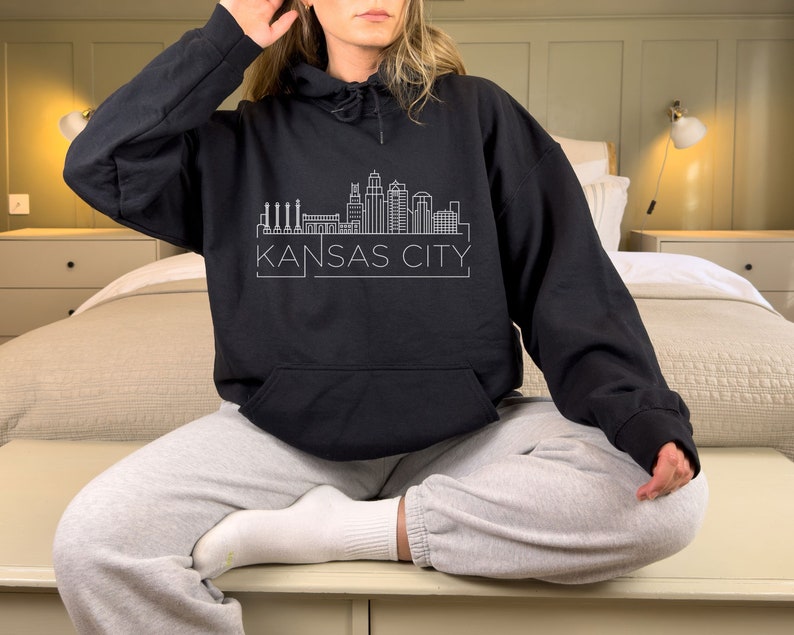 Może przedstawiać: Czarna bluza z kapturem z białym obrysem panoramy Kansas City i nazwą miasta wydrukowaną poniżej. Bluza ma przednią kieszeń i kaptur ze sznurkiem. Modelka ma na sobie szare spodnie dresowe.