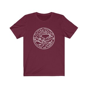 May include: A maroon t-shirt with a white graphic of the state seal of Tennessee. The seal features a mountain range, a river, and the words "Tennessee, Agriculture and Commerce".