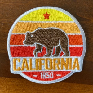 May include: Embroidered patch with a brown bear silhouette on a sunset-colored background. The patch says "CALIFORNIA - 1850 -" in yellow and white lettering.