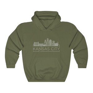 Może przedstawiać: Oliwkowo-zielona bluza z kapturem z białym rysunkiem linii horyzontu Kansas City. Tekst "KANSAS CITY" jest wydrukowany na biało pod linią horyzontu. Bluza ma przednią kieszeń i kaptur ze sznurkiem.