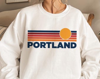May include: White sweatshirt featuring a retro sunset graphic and the word "PORTLAND" in blue lettering.