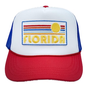 May include: A white and red trucker hat with a blue mesh back. The front of the hat features a yellow embroidered patch with the words "FLORIDA" and a stylized sun with orange, red, and blue stripes.