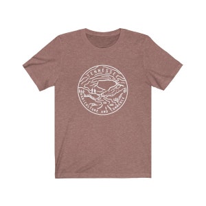 May include: A heather brown t-shirt with a white graphic of the Tennessee state seal. The seal features a mountain range, a river, and the words "Tennessee, Agriculture and Commerce".