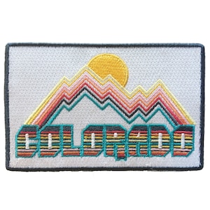 May include: A white embroidered patch with a colorful mountain range and a yellow sun. The word "COLORADO" is embroidered in colorful stripes below the mountains.