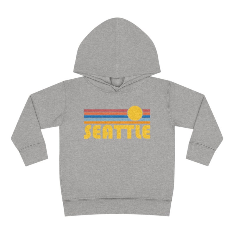 Pode incluir: Camisola de moletom cinza com capuz com um gr&aacute;fico retr&ocirc; de um p&ocirc;r do sol sobre um horizonte urbano com a palavra "SEATTLE" em amarelo.