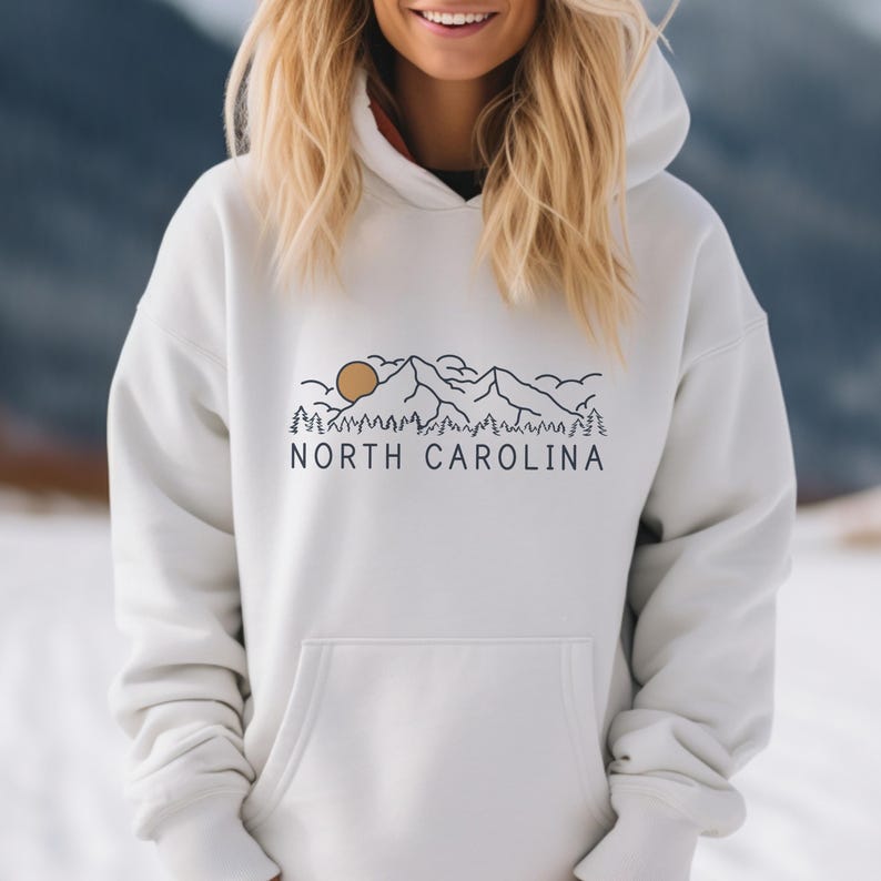 May include: White hooded sweatshirt with a graphic design of mountains, trees, and a sun with the text "NORTH CAROLINA" printed on the front.
