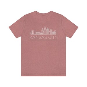 May include: A heather rose colored t-shirt with a white outline of the Kansas City skyline and the text "KANSAS CITY" in a white box.