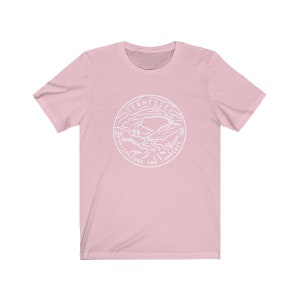 May include: A pink t-shirt with a white graphic of the state seal of Tennessee. The seal features a mountain landscape with a river flowing through it. The text "Tennessee Agriculture and Commerce" is inscribed around the seal.