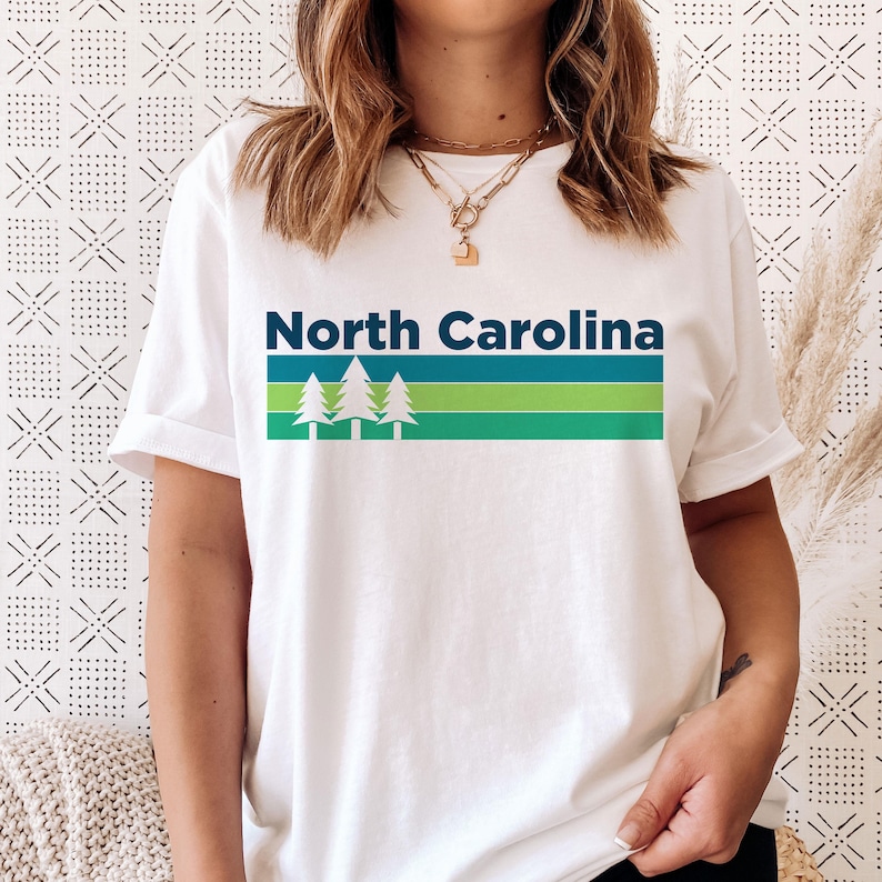 Pode incluir: Camiseta branca com um design retro azul e verde que diz "North Carolina" com tr&ecirc;s pinheiros.