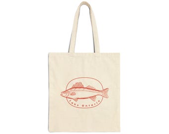 Puede incluir: Bolso tote de color crema con una ilustración roja de un pez dentro de un círculo, con las palabras "Lake Ontario" alrededor del pez. El bolso tiene dos asas largas.