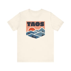May include: A white t-shirt with a graphic print of a mountain range with a red sun and the word "TAOS" in black.