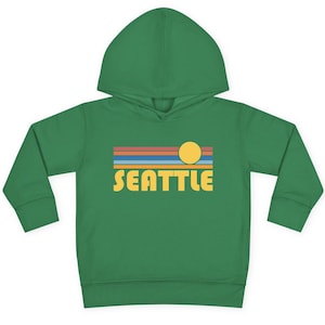 Pode incluir: Uma jaqueta de moletom verde para crian&ccedil;as pequenas com um gr&aacute;fico de p&ocirc;r do sol retr&ocirc; e a palavra "SEATTLE" em letras amarelas.