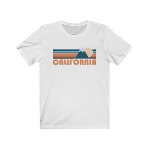 May include: White t-shirt with a retro-style graphic of a mountain range, sun, and the word "CALIFORNIA" in orange.