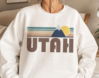 May include: White sweatshirt featuring a brown graphic of a mountain range and the word "UTAH" in brown lettering. The graphic is set against a backdrop of colourful stripes.