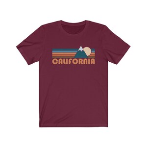 May include: A maroon short-sleeved t-shirt with a retro-style graphic of a mountain range, sun, and the word "CALIFORNIA" in orange.