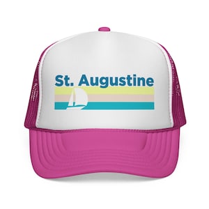 May include: A pink and white trucker hat with the text "St. Augustine" in blue. The hat features a sailboat graphic and horizontal stripes in yellow, beige, and teal. The brim and mesh backing are pink.