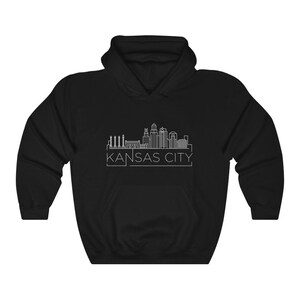 Może przedstawiać: Czarna bluza z kapturem z białym rysunkiem linii panoramy Kansas City. Nazwa miasta jest wydrukowana na dole na biało. Bluza ma przednią kieszeń i kaptur ze sznurkiem.